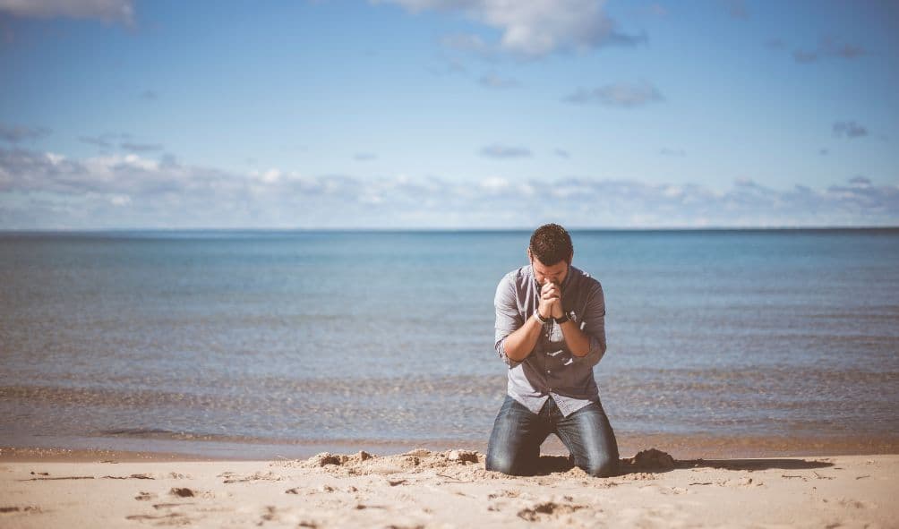 homem orando de joelho à beira da praia